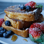 French Toast topped with blueberries and strawberries, drizzled with syrup and dusted with powdered sugar, on a grey plate.