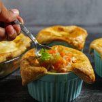 Chicken Pot Pie with Puff Pastry in a blue ramekin. A spoon with the contents of the pot pie, carrots, chicken, celery and potatoes, is digging into the pie.