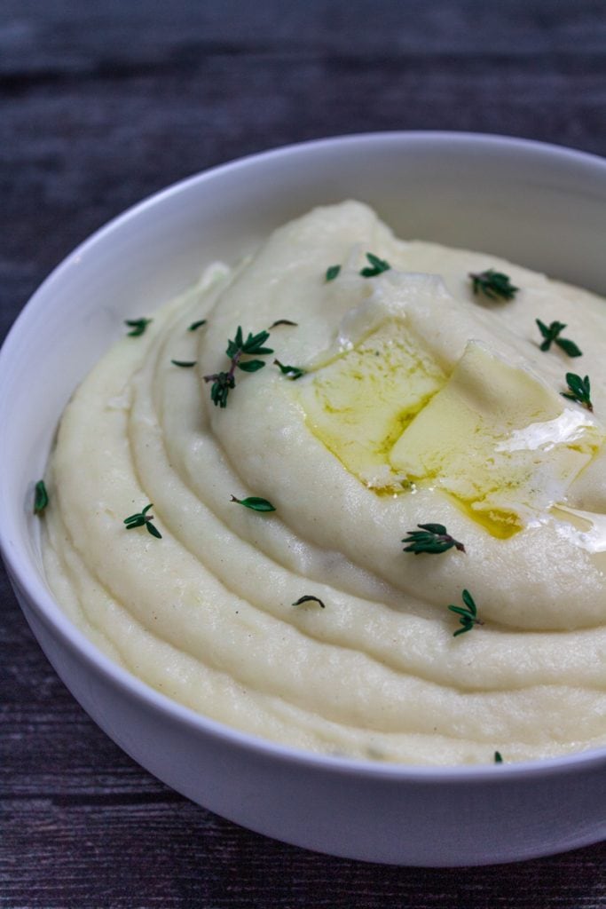 Bowl of Creamy Garlic and Herb Mashed Potatoes, topped with fresh thyme leaves and two pats of butter.