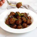 Honey Glazed Garlic And Ginger Meatballs garnished with thinly sliced scallions in a white bowl. One meatball is being picked up with a gold fork.