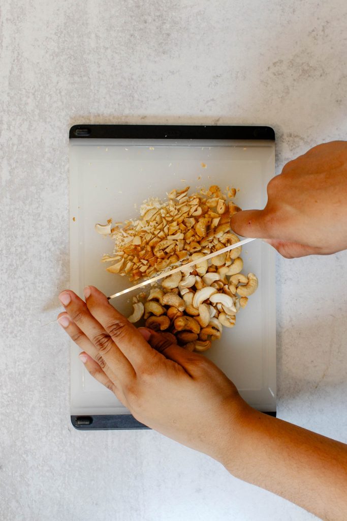 Rough chopping roasted cashews on a plastic cutting board with a chefs knife.