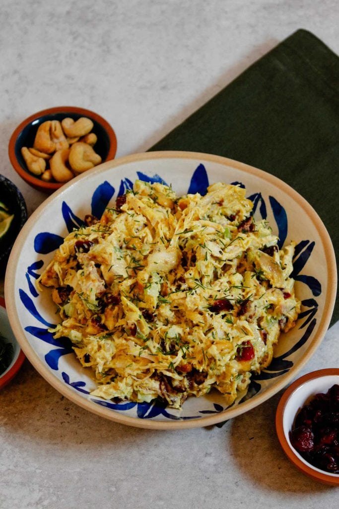 Curry chicken salad plated in white and blue bowl, surrounded by small bowls of cashes, lime wedges, dill, and dried cranberries.