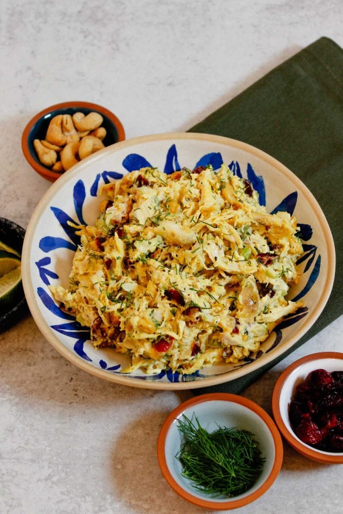 Curry chicken salad plated in white and blue bowl, surrounded by small bowls of cashes, lime wedges, dill, and dried cranberries.