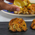 Squeezing a lemon onto Maryland crab cakes, placed on a black plate.