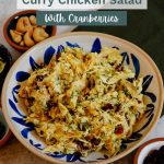 Curry chicken salad plated in white and blue bowl, surrounded by small bowls of cashes, lime wedges, dill, and dried cranberries.