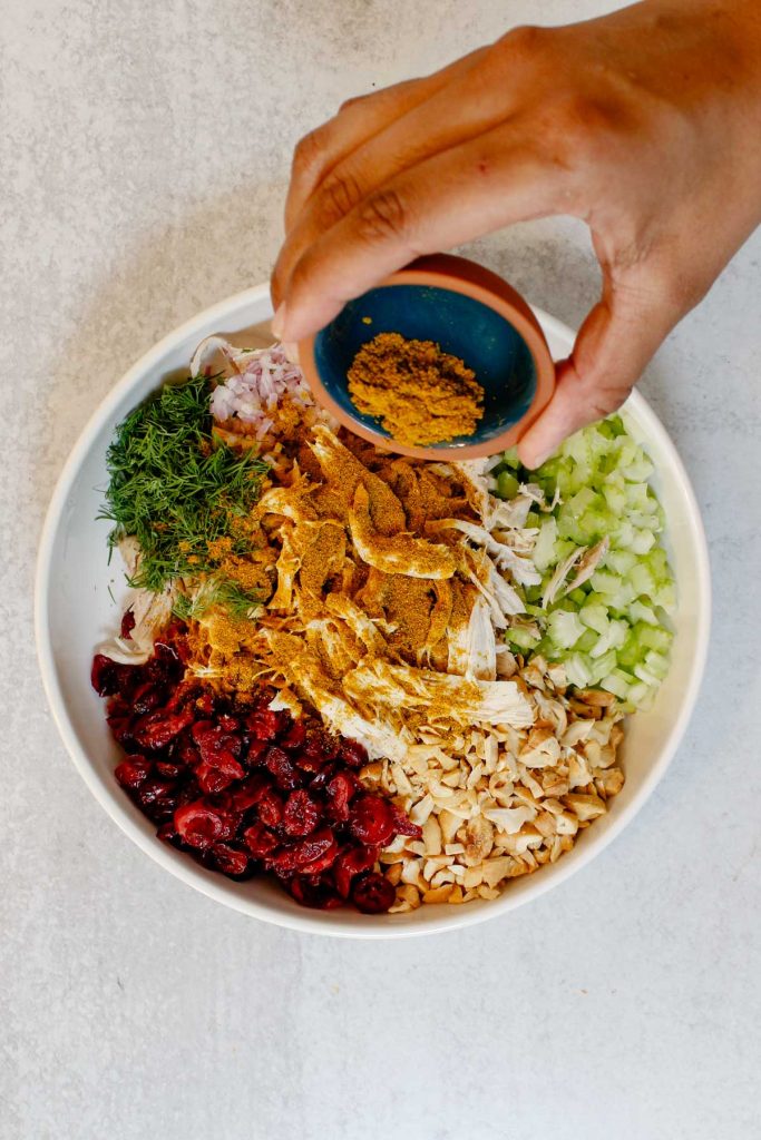 Sprinkling curry powder onto a bowl filled with curry chicken salad ingredients.