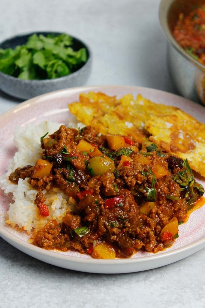 Puerto Rican picadillo plated with white rice and tostones on a pick plate, next to a large skillet filled with picadillo.