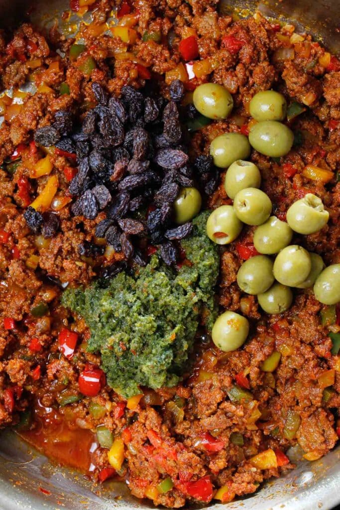 Olives, raisins, and sofrito added to picadillo in a skillet.