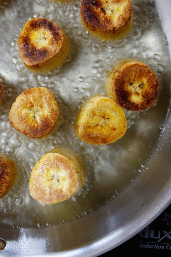 Golden brown green plantains frying in a large skillet with vegetable oil.