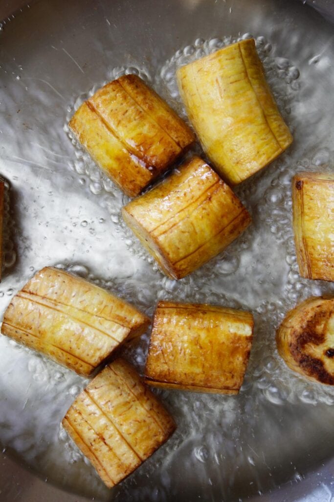 Golden brown green plantains frying in a large skillet with vegetable oil.