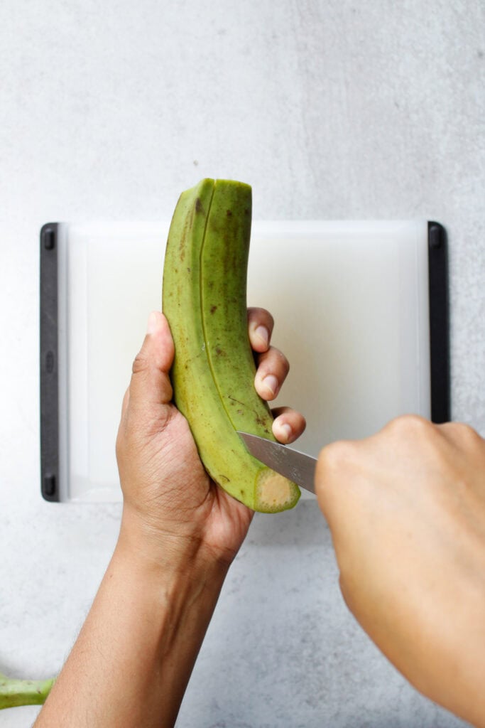 Slicing shallow cuts down the length of a green plantain using a paring knife.