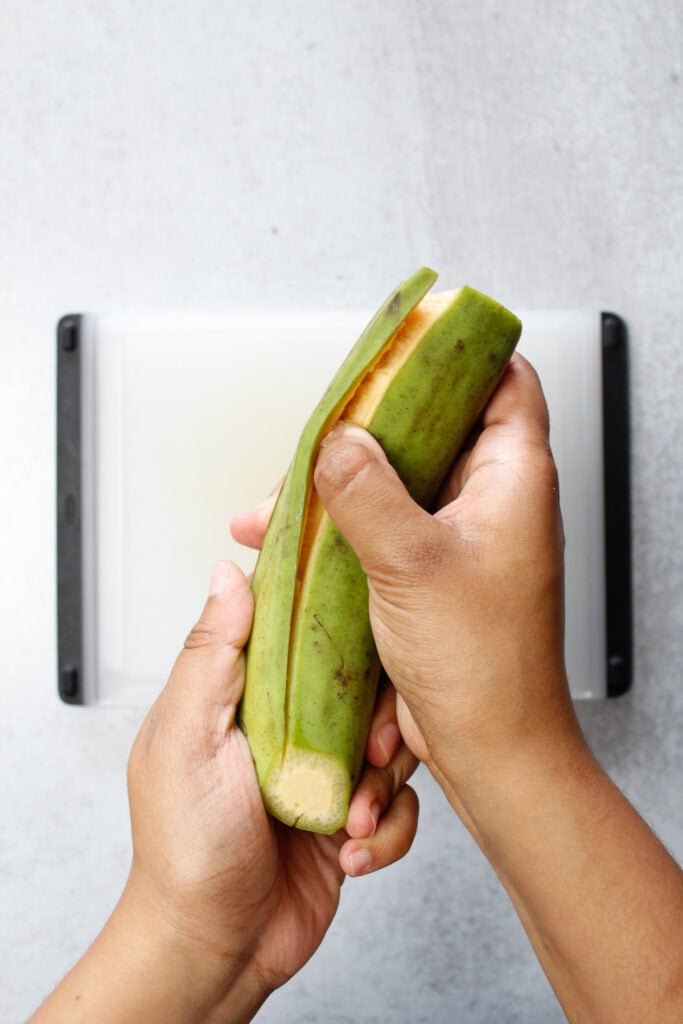 Sliding my thumb under the skin of a green plantain to remove the peel.