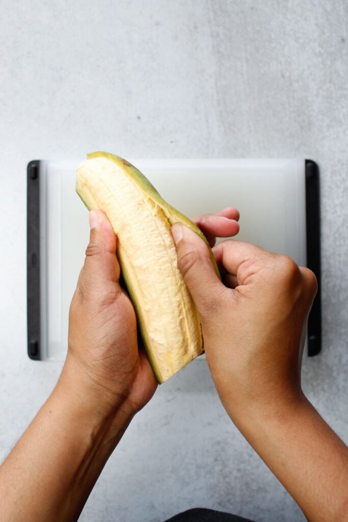 Sliding my thumb under the skin of a green plantain to remove the peel.