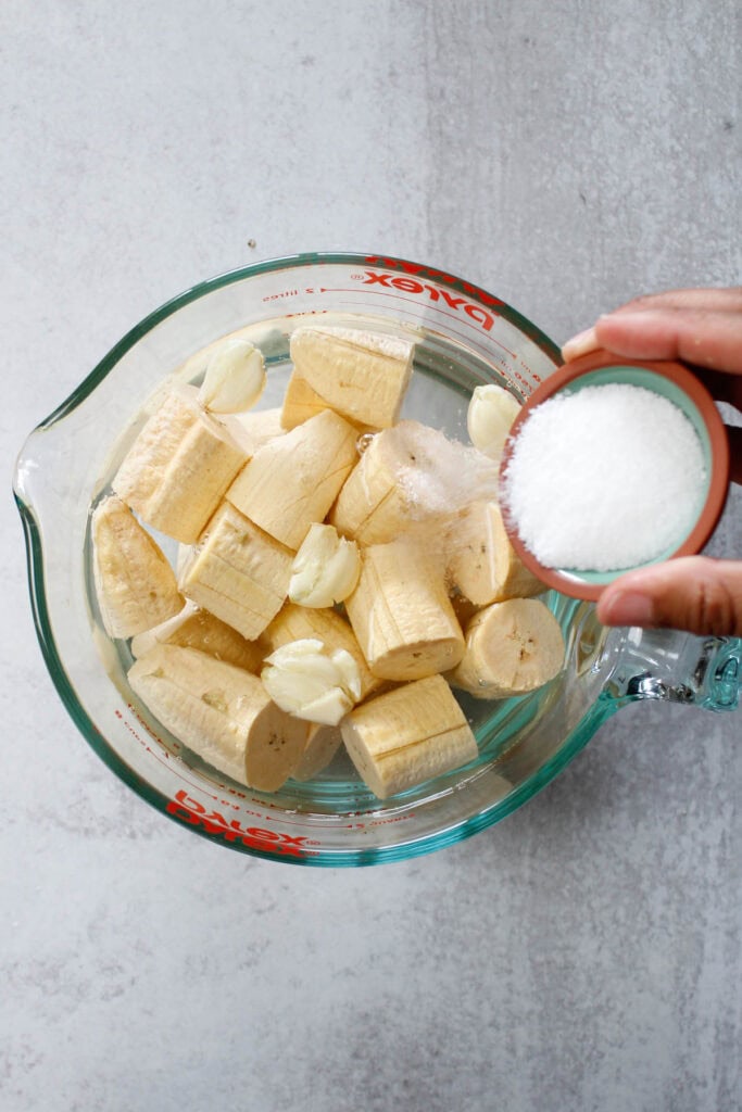 Pouring salt into a large pyrex measuring cup with smashed garlic cloves and sliced green plantains.