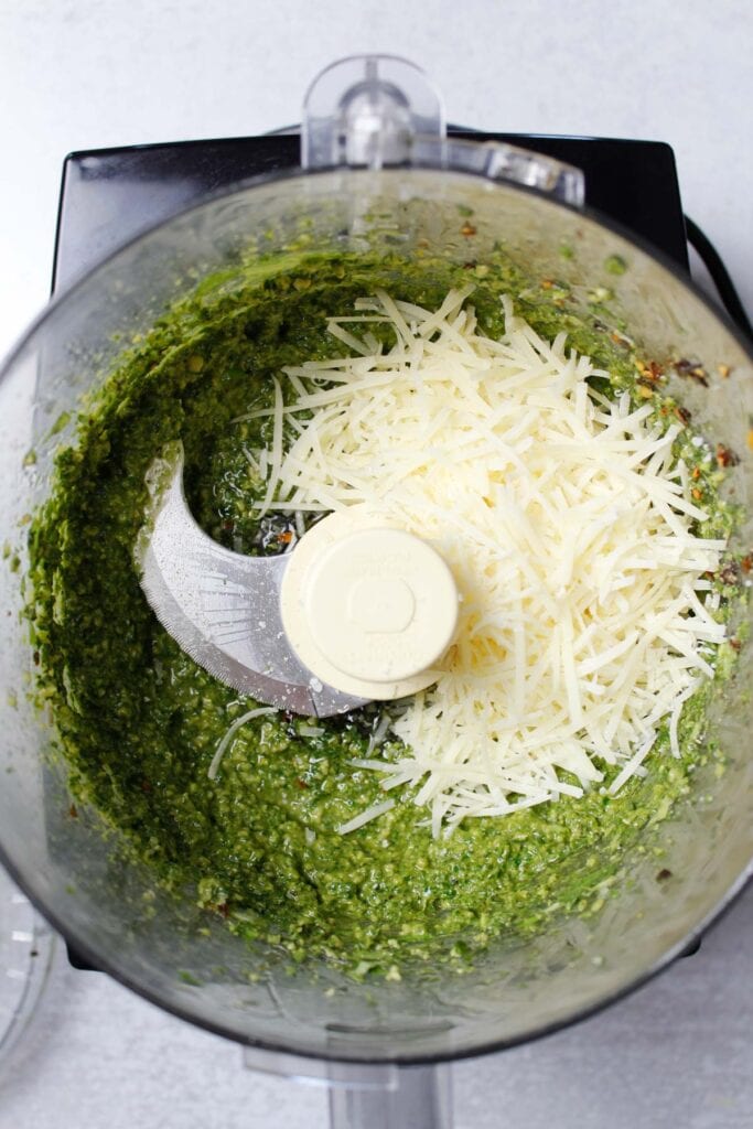 Adding cheese to pesto sauce in a food processor.