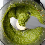 Spatula showing off some basil pesto sauce in front of a food processor filled with the same pesto.