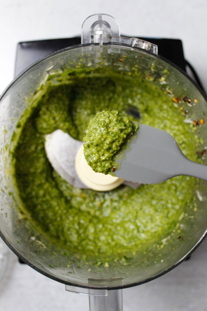 Spatula showing off some basil pesto sauce in front of  a food processor filled with the same pesto.