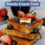 A stack of Brioche French Toast being drizzled with syrup, with strawberries, blueberries and a pat of butter.