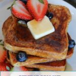 A stack of Brioche French Toast drizzled with syrup, with strawberries, blueberries and a pat of butter.