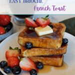 A stack of Brioche French Toast drizzled with syrup, with strawberries, blueberries and a pat of butter.