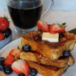 A stack of Brioche French Toast being drizzled with syrup, with strawberries, blueberries and a pat of butter.