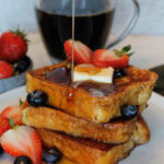 A stack of Brioche French Toast being drizzled with syrup, with strawberries, blueberries and a pat of butter.