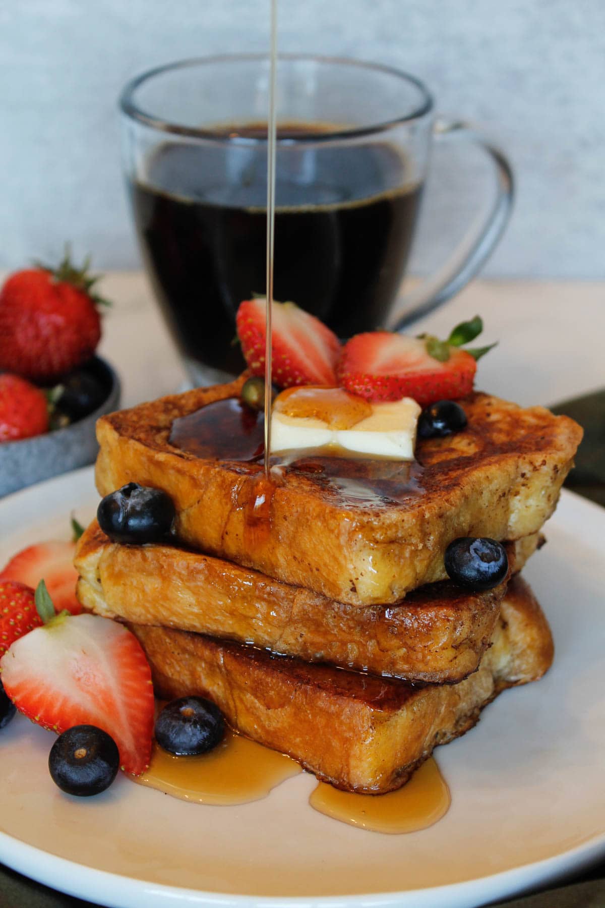 A stack of Brioche French Toast being drizzled with syrup, with strawberries, blueberries and a pat of butter.