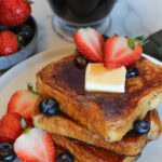 A stack of Brioche French Toast drizzled with syrup, with strawberries, blueberries and a pat of butter.