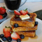 A stack of Brioche French Toast drizzled with syrup, with strawberries, blueberries and a pat of butter.