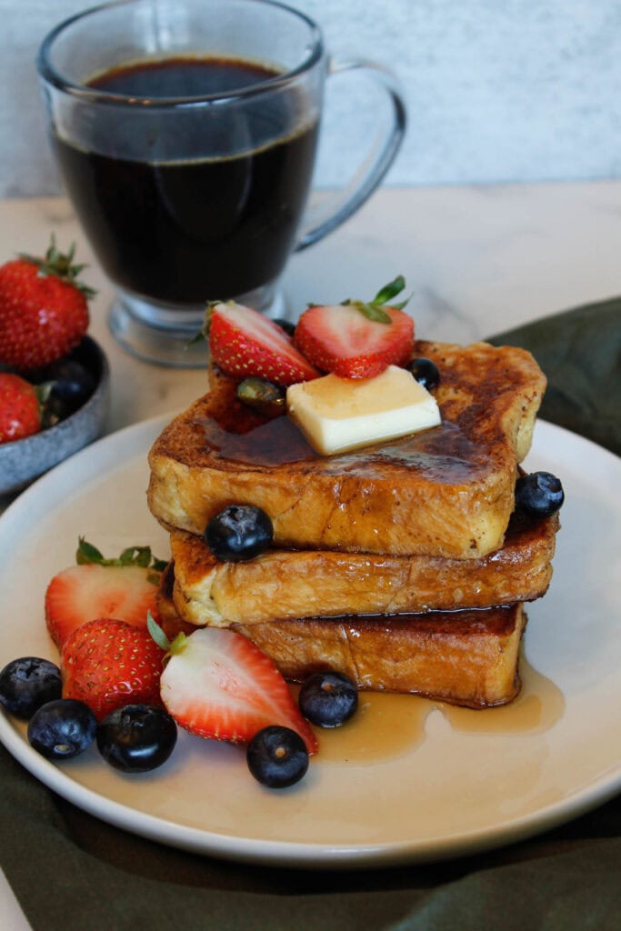 A stack of Brioche French Toast drizzled with syrup, with strawberries, blueberries and a pat of butter.