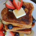 A stack of Brioche French Toast drizzled with syrup, with strawberries, blueberries and a pat of butter.