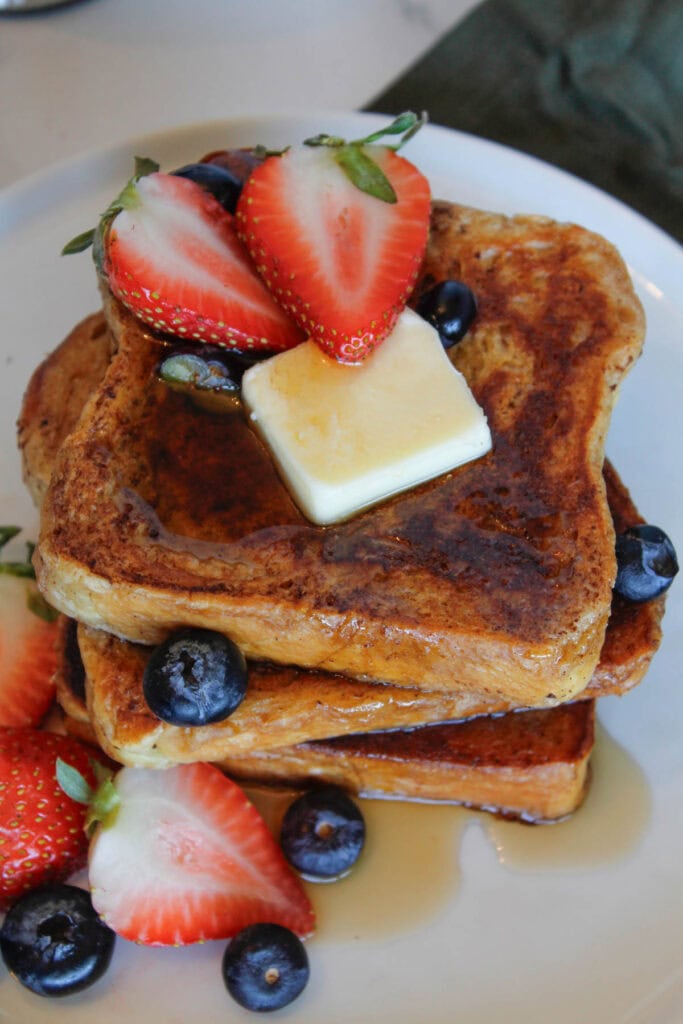 A stack of Brioche French Toast drizzled with syrup, with strawberries, blueberries and a pat of butter.