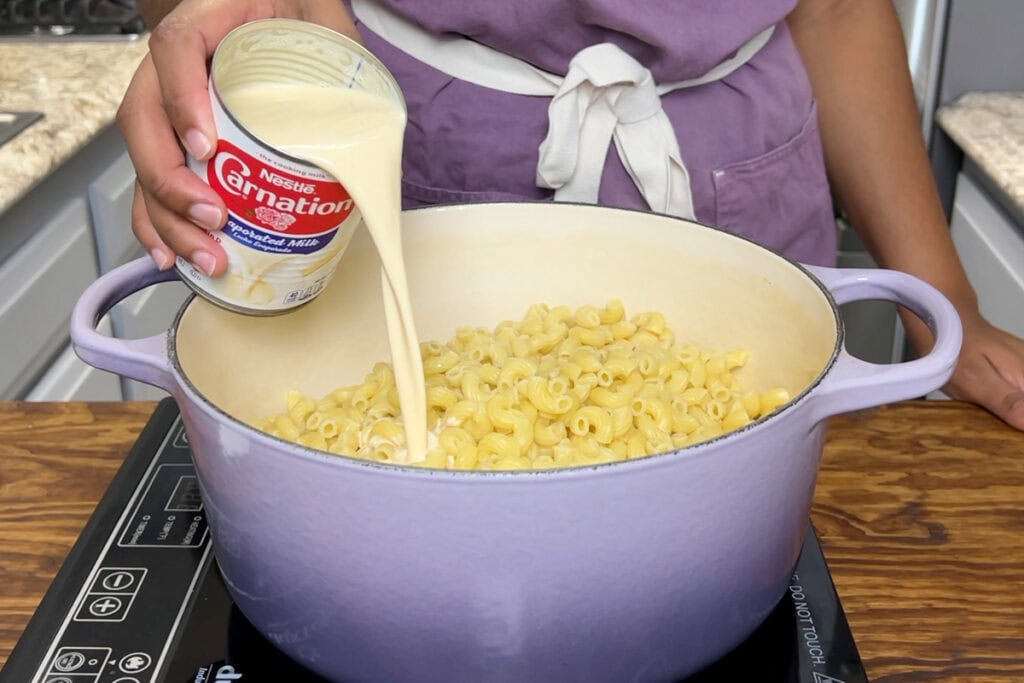 Pouring evaporated milk in elbow noodles for baked macaroni and cheese.